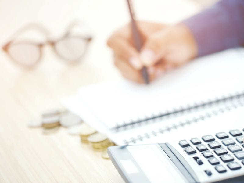 persona fa i conti con la calcolatrice, sullo sfondo un paio di occhiali e dei soldi in moneta